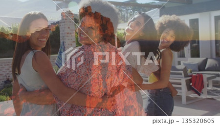 Smiling four friends linking arms on backyard patio, with white lounge chairs, red towel and plants 135320605