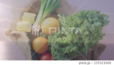 Burlap sack lined with green cloth resting on wooden dock by calm water, with fresh vegetables 135321008
