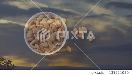 Reflecting white ceramic bowl holding almonds on reflective glass table at dusk, showing cloudy sky Reflecting white ceramic bowl holding almonds on reflective glass table at dusk, showing cloudy sky 135321020