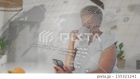 Holding smartphone and coffee mug, woman analyzing world map projection in home kitchen with plants 135321241