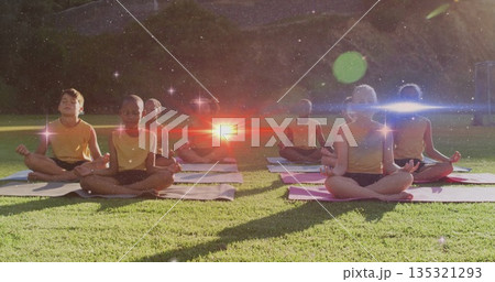 Meditating children sitting cross-legged on yoga mats in park on grassy lawn, wearing yellow tops 135321293