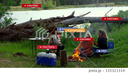 Gathering friends sitting around campfire on lakeshore with fallen log checking phone near cooler Gathering friends sitting around campfire on lakeshore with fallen log checking phone near cooler 135321314