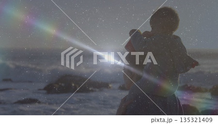 Standing mother holding toddler on rocky shoreline at sunset, wearing jackets, copy space 135321409