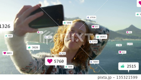 Posing woman wearing sweater holding smartphone near lakeside, with floating social media icons 135321599