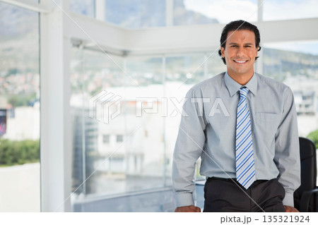 Mid adult man sitting at desk near floor-to-ceiling windows in modern office workspace, copy space Mid adult man sitting at desk near floor-to-ceiling windows in modern office workspace, copy space 135321924