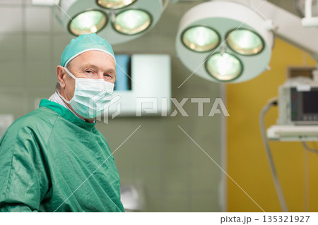 Senior male surgeon wearing green scrubs and mask standing under surgical lights in operating room 135321927
