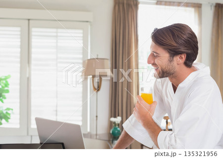 Adult male leaning on table in living room holding orange juice while browsing laptop, copy space Adult male leaning on table in living room holding orange juice while browsing laptop, copy space 135321956