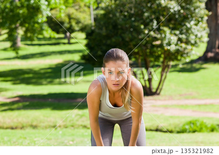 Woman pausing leaning forward with hands on knees in sunlit grassy park wearing sportswear 135321982