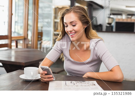 Woman sitting at wooden table inside cafe wearing gray top smiling at smartphone beside coffee cup Woman sitting at wooden table inside cafe wearing gray top smiling at smartphone beside coffee cup 135321986