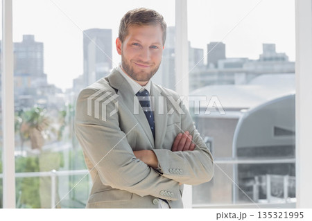 Businessman standing with arms crossed in office by floor-to-ceiling window showing city skyline 135321995