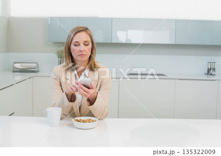 Senior woman sitting at modern kitchen island holding smartphone beside cereal bowl with milk 135322009