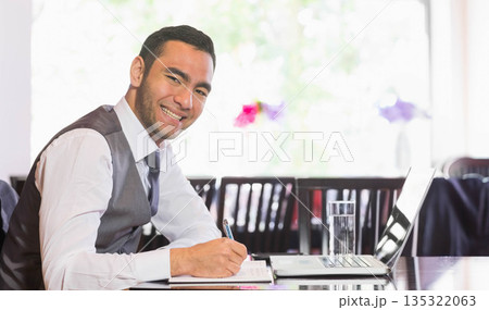 Man in business attire taking notes at cafe table with laptop and notebook, copy space 135322063