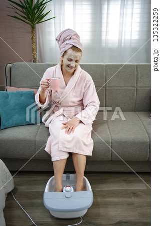 Woman relaxing on sofa with face mask and enjoying a foot bath while drinking tea 135322259