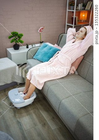 Woman relaxing on sofa with face mask and enjoying feet bath at home 135322260