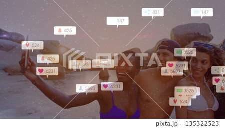 Smiling four friends posing for group selfie on sandy beach at sunset with smartphone notifications 135322523