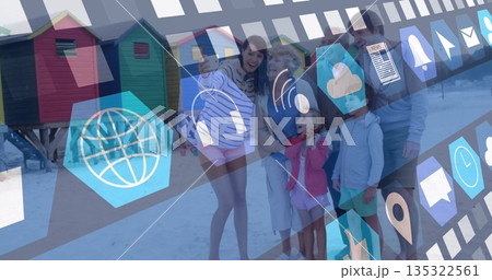 Smiling extended family posing at shoreline, with smartphone, colorful beach huts and digital icons 135322561