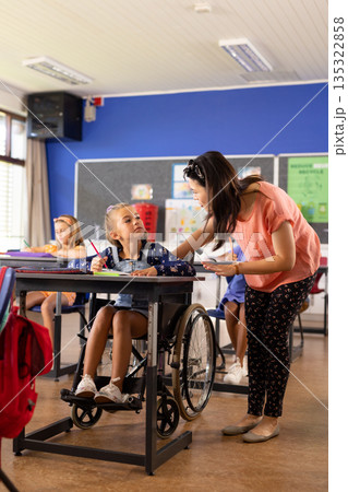 Asian teacher assists a girl in a wheelchair at a school classroom desk Asian teacher assists a girl in a wheelchair at a school classroom desk 135322858