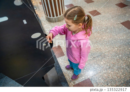 Little girl collecting coins from money exchange machine in shopping mall kids area. Preparation for play, childhood independence moment and family leisure routine in real life commercial environment Little girl collecting coins from money exchange machine in shopping mall kids area. Preparation for play, childhood independence moment and family leisure routine in real life commercial environment 135322917