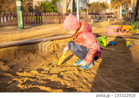 Little girl playing with sand in sandbox at playground during spring. Outdoor childhood activity, creative play and everyday life in urban park 135322918