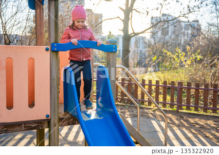 Little girl sliding down playground slide in city park during spring. Childhood outdoor activity, seasonal play and everyday leisure moment in real life urban environment Little girl sliding down playground slide in city park during spring. Childhood outdoor activity, seasonal play and everyday leisure moment in real life urban environment 135322920