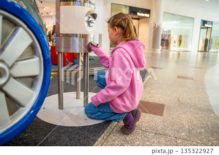 Little girl standing near gemstone vending machine and waiting for random stone in shopping mall kids area. Childhood curiosity moment, family leisure experience in real life commercial environment 135322927