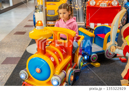 Little girl riding children train attraction in shopping mall kids area. Childhood entertainment activity, playful movement and family leisure time in real life commercial environment 135322929