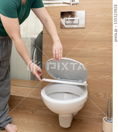 Person's hands lifting white toilet seat lid in a contemporary bathroom setting, focusing on hygiene and domestic routine 135323928