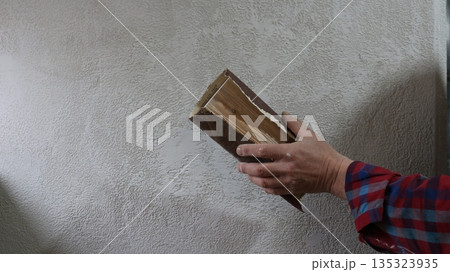 A man's hand is shown using a homemade device made of a small wooden board and sandpaper to clean and smooth plaster on a room wall; the DIY sanding tool is in the man's hand, scraping the plaster A man's hand is shown using a homemade device made of a small wooden board and sandpaper to clean and smooth plaster on a room wall; the DIY sanding tool is in the man's hand, scraping the plaster 135323935