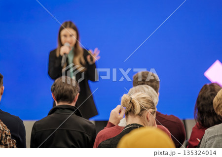 Speaker delivers talk to engaged audience during presentation with blue backdrop and microphone Speaker delivers talk to engaged audience during presentation with blue backdrop and microphone 135324014