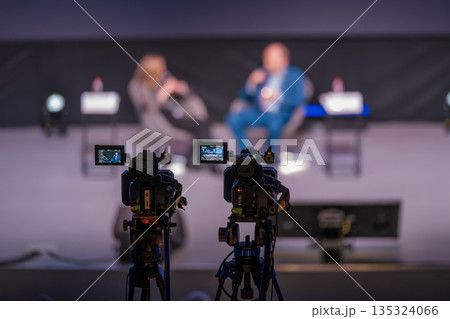 Two cameras capture live panel discussion with blurred speakers on stage and studio setup for a professional broadcast Two cameras capture live panel discussion with blurred speakers on stage and studio setup for a professional broadcast 135324066
