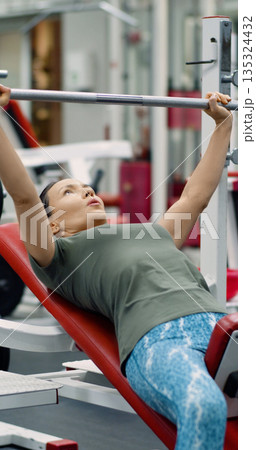 Gym workout - with determined focus, a middle-aged woman completes her hammer machine bench press routine, building upper body strength and enhancing fitness. Gym workout - with determined focus, a middle-aged woman completes her hammer machine bench press routine, building upper body strength and enhancing fitness. 135324432