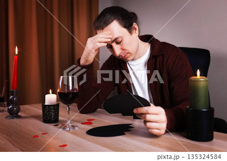 Sad man holding a broken black heart at a table with wine and candles. Upset lonely guy suffering from breakup or divorce. Anti-Valentine's Day concept. High quality photo Sad man holding a broken black heart at a table with wine and candles. Upset lonely guy suffering from breakup or divorce. Anti-Valentine's Day concept. High quality photo 135324584