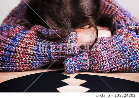Depressed woman hiding face in arms near a broken black heart. Sad lonely female suffering from a breakup. Anti-Valentine's Day concept. High quality photo 135324802