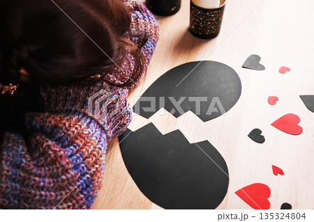 Depressed woman lying head down on a table next to a broken black heart. Sad lonely female suffering from a breakup. Anti-Valentine's Day concept. High quality photo 135324804