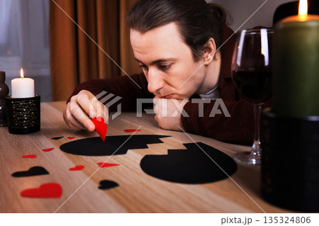 Sad lonely man gazing at a broken black heart on a table with wine. Upset guy suffering from breakup. Anti-Valentine's Day concept. High quality photo Sad lonely man gazing at a broken black heart on a table with wine. Upset guy suffering from breakup. Anti-Valentine's Day concept. High quality photo 135324806