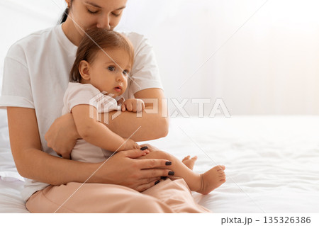 Caucasian mother holding baby close while sitting on bed, showing emotional connection 135326386