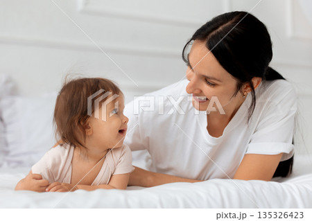 Mother and infant baby lying together on bed at home and smiling Mother and infant baby lying together on bed at home and smiling 135326423