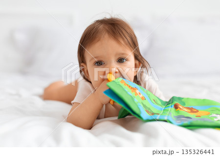 Baby lying on a bed and chewing a soft fabric book Baby lying on a bed and chewing a soft fabric book 135326441