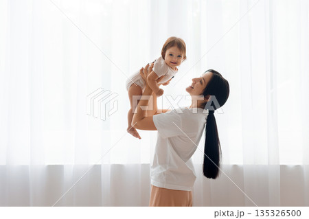 Smiling mother lifting her infant baby near window at home 135326500