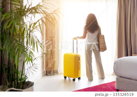 Rear view of woman standing in hotel room near window with suitcase Rear view of woman standing in hotel room near window with suitcase 135326519