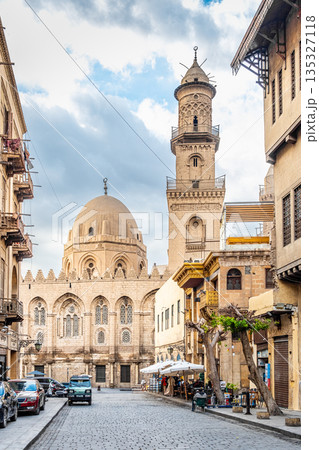 Historic Qalawun Complex in Cairo, featuring intricate architecture, tall minarets, and domes under a blue sky with clouds. A significant cultural landmark on Al-Muizz Street. 135327118