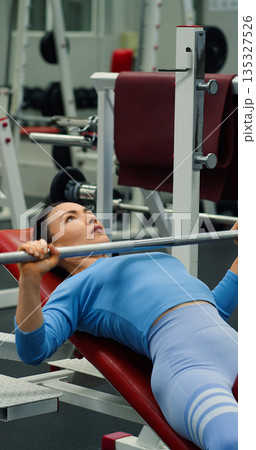 Gym workout. Bench press. Middle aged Caucasian woman trains her pectoral muscles in the gym. Caucasian woman trains chest muscles using bench press during gym workout. 135327526