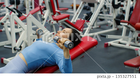 A middle aged fit Caucasian woman during gym workout. Bench press with dumbbells for chest. Strength training with bench press highlights a woman's determination and focus. A middle aged fit Caucasian woman during gym workout. Bench press with dumbbells for chest. Strength training with bench press highlights a woman's determination and focus. 135327551