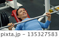 Gym workout. Bench press. Caucasian woman trains her pectoral muscles in the gym. Caucasian woman trains chest muscles using bench press during gym workout. Close-up photo. 135327583