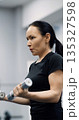 Close-up vertical photo of a fit middle aged woman training biceps with dumbbells during an intense gym workout. Biceps with dumbbells workout. Cropped footage of a working out woman. 135327598
