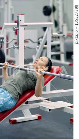 Gym workout. Photo of a middle-aged woman pushes through a bench press set on the hammer machine, combining steady breathing and firm control for an effective workout. Gym workout. Photo of a middle-aged woman pushes through a bench press set on the hammer machine, combining steady breathing and firm control for an effective workout. 135327599