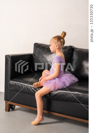 Young dancer with a cochlear implant sits in profile on a sofa wearing a lavender tutu. Inclusive performance, hearing support, and thoughtful childhood through ballet and quiet focus. 135330730