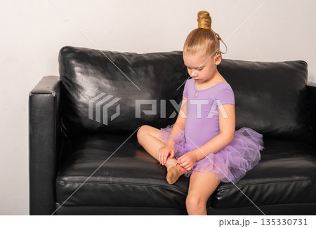 Young ballerina with a cochlear implant sits on a black couch during a quiet rehearsal break. Inclusive ballet, hearing technology, and focused childhood through rest and reflection. 135330731
