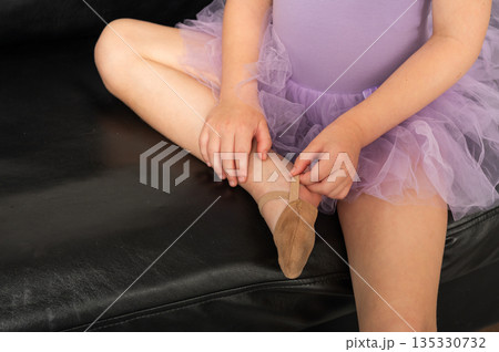 A close view of a young dancer adjusting her ballet shoe on a sofa. Inclusive training, hearing support, and careful preparation through dance and discipline. 135330732