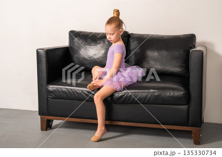 A young ballerina with a cochlear implant sits quietly on a black couch after rehearsal. Inclusive ballet, hearing technology, and focused childhood through rest and recovery. 135330734